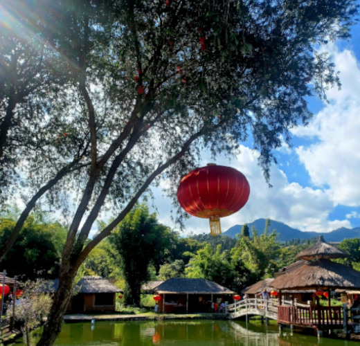 Red lantern hanging over a green pond in Pai, North Thailand. A a wooden bridge and bamboo huts are framed by trees and mountains under a blue sky