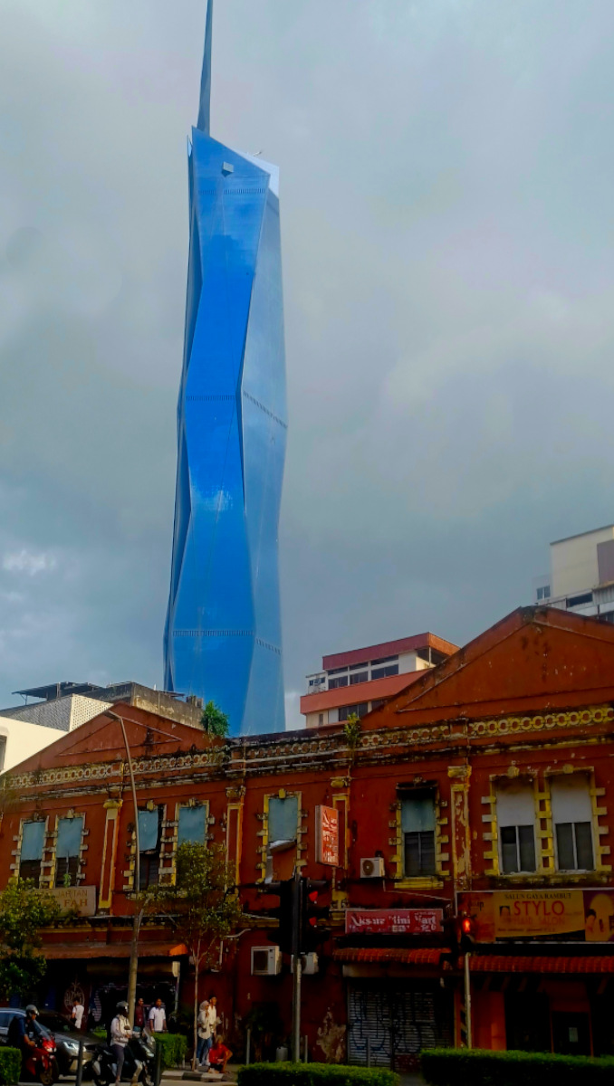 Merdeka 118 skyscraper in Chinatown, Kuala Lumpur, towering over brick red colonial buildings under a grey, cloudy sky, showcasing its bright blue and shiny facade