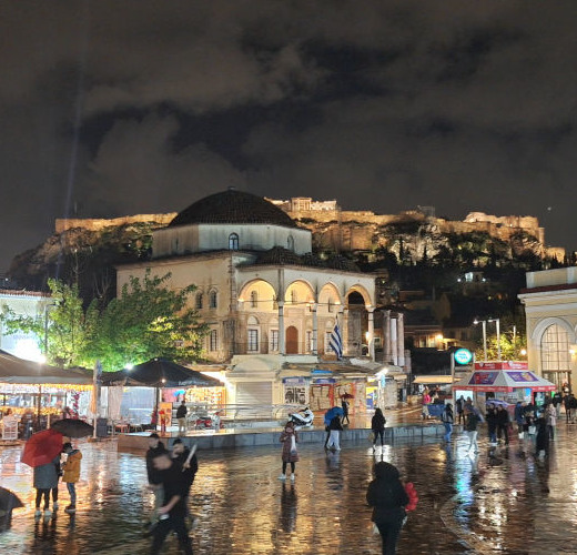 Shining cobbled streets of Monastiraki Square in Athens at night with the Acropolis against a dark sky.